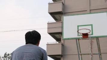 hombre asiático en ropa deportiva jugando baloncesto en el patio de recreo al aire libre. los jugadores masculinos de baloncesto practican al aire libre en las canchas locales. video