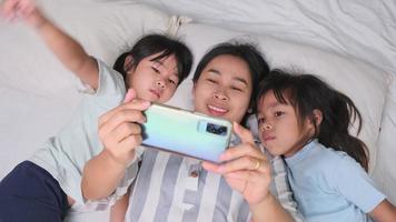 Happy Asian family enjoying with smartphone at cozy home. Smiling mother and cute daughters using phone, Take a selfie or video clip to remember in bed at home. Good time at home