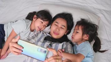 feliz família asiática desfrutando com smartphone em casa aconchegante. mãe sorridente e filhas fofas usando telefone, tire uma selfie ou videoclipe para lembrar na cama em casa. bom tempo em casa