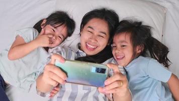 famille asiatique heureuse appréciant avec smartphone dans une maison confortable. mère souriante et filles mignonnes utilisant le téléphone, prenez un selfie ou un clip vidéo à retenir au lit à la maison. bon moment à la maison video