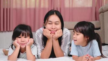 dos tiernas hijas besando a feliz madre. niños felices jugando con su madre acostada en una cama en casa y mirando la cámara. video