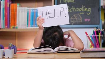 Asian little girl feeling sad and tired sitting at the table with many books and holding a paper with word Help. Learning difficulties. Home schooling. video