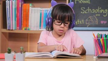 menina bonitinha com fones de ouvido ouvindo audiolivros e olhando livros de aprendizagem de inglês em cima da mesa. aprendendo inglês e educação moderna video