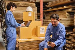 dos trabajadores y colegas de uniformes de seguridad usan computadoras portátiles y teléfonos móviles para verificar las existencias de pedidos de envío en el almacén de paquetes, fábrica de fabricación de papel para la industria del embalaje, servicio de transporte logístico. foto