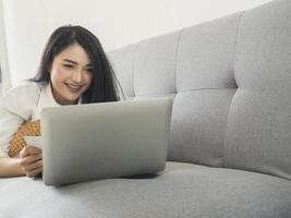 hermosa mujer con un vestido blanco, jugando una computadora portátil en el sofá. ideas de negocios y compras en linea foto