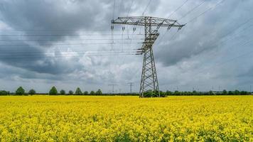 Beautiful farm landscape with yellow rapeseed at blossom field, wind turbines to produce green energy and high voltage power lines in Germany, at Spring and dramatic rainy sky. photo