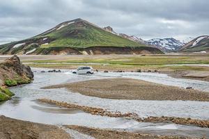 A car is fording a mountain creek at surreal magic Icelandic landscape of colorful rainbow volcanic Landmannalaugar mountains, near a biggest camping site there with tourists and hikers, Iceland photo