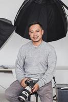 fotógrafo asiático sonriente sentado y sosteniendo la cámara en el estudio foto