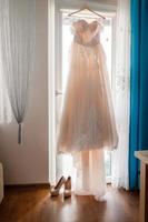 preparación de la mañana de la novia. hermoso vestido largo blanco de boda y zapatos de colinas altas en el fondo de una gran ventana en casa. día de la boda foto