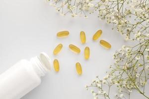 Oil yellow gelatin capsules with white bottle on white background with flowers, vitamins and antioxidant concept Omega 3, liver cod or evening primose oil for healthcare. Minimalism. Copy space. photo