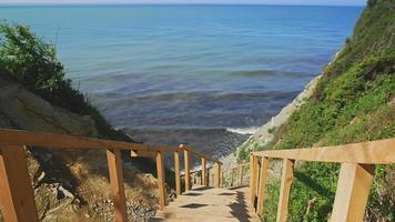 Brown wooden stairs with handrails leading to beach between hills with grass against endless blue sea waves rolling on coastline video