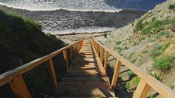 Brown wooden stairs with handrails on hilly coastline lead to blue sea waves rolling on pebble beach reflecting bright sunlight video