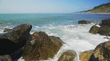 Las olas del océano espumoso blanco azul chocan contra grandes piedras marrones contra la costa montañosa con bosques densos verdes en cámara lenta de verano video