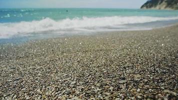 Foaming sea waves roll on grey brown pebble beach against hilly coastline under bright sunlight on summer day slow motion