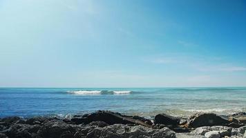 Dense white foam appears on large endless blue sea wave with grey wet stones on foreground under bright sunlight slow motion video