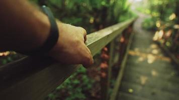 Guy traveler walks along brown wooden bridge and touches handrails with hand with black smartwatch on summer day close view video