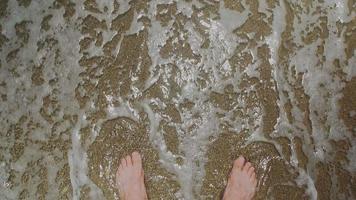 Sea transparent wave with foamy tip rolls and washes guy tourist bare feet standing on wet yellow sandy beach first point view video