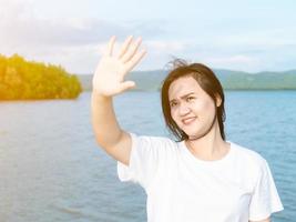 chica asiática con camisa blanca sonriendo pon tus manos para bloquear la luz del sol de los ojos que bajan a tus ojos. en la sesión de fotos de medio cuerpo del mar soleado de vacaciones. obtener el poder de la naturaleza.