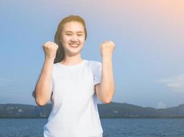 chica asiática con camisa blanca con ambas manos, hazlo si estás feliz. que logro el objetivo se ve feliz desafiar el viento en el mar y el sol el dia que salio de vacaciones foto