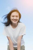 una chica asiática con una camisa blanca se inclina y la saluda con una sonrisa. se veía feliz, siendo soplada con fuerza, desafiando el viento en el mar y el sol. el día que ella salió de vacaciones foto