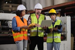 Safety uniform workers and industrial engineers with hardhat use laptop computer to check and control machines. Three professionals work in paper manufacturing factory, maintain production equipment. photo