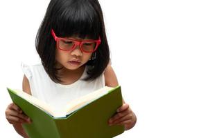 Happy Asian little preschool girl wearing red glasses holding and read a green book on white isolated background. Concept of school kid and education in elementary and preschool, home school photo