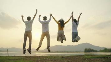 vier mensen groep gelukkige tieners die handen opsteken die op zonsondergangberg en rivierachtergrond springen. video