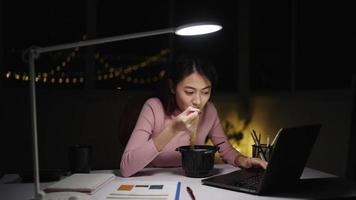 mujer independiente asiática usa ropa rosa comiendo fideos preparados mientras trabaja demasiado. escriba con la computadora portátil en el escritorio y la lámpara en casa por la noche. concepto de trabajo independiente de las personas con el tiempo.