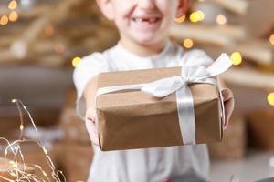 simple gift in kraft paper packaging tied with a white ribbon in the hands of a child boy in a t shirt. baby holding gift box on christmas background. concept of new year, mothers day, Valentines day photo