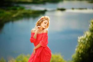 beautiful young blonde woman with a flower wreath on her head. Beauty girl with flowers hairstyle in a red long dress near the lake. Fashion photo, copy space photo