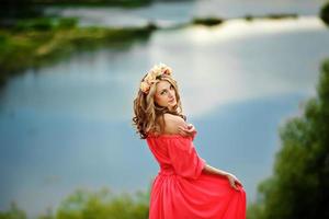 beautiful young blonde woman with a flower wreath on her head. Beauty girl with flowers hairstyle in a red long dress near the lake. Fashion photo, copy space photo