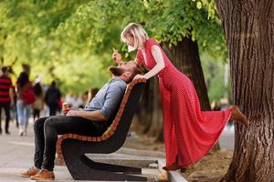 smiling hipsters couple having fun and eating ice cream in the city. stylish young man with beard is sitting on a wooden bench and blonde woman in red dress woman fools around and plays with him photo