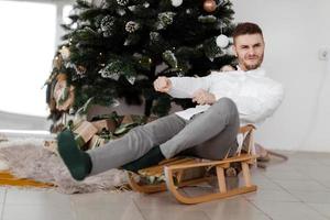 cheerful man sitting on sleigh at home near Christmas tree. man on a sleigh. Christmas good mood. Family and holiday concept. photo