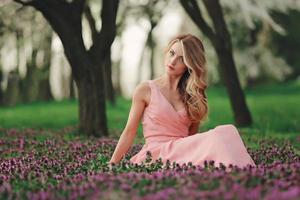 hermosa joven rubia en flores de colores. chica con maquillaje y peinado en vestido rosa en el floreciente parque de primavera. día de la Mujer foto