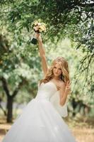 Cute young bride with long hairs holding her wedding bouquet includes white roses and other flowers. Beautiful white marriage dress. Pretty girl on green trees background. photo