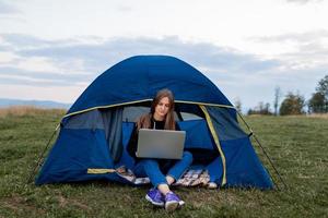 Girl with laptop in the mountains. Tourist woman uses a laptop in nature, freelancer typing on a computer near a tent in a journey against the backdrop of a mountain landscape