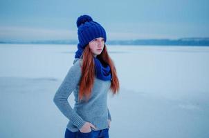 Portrait of young red hair girl with freckles wearing at blue knitted wool hat and scarf in winter day background ice. photo