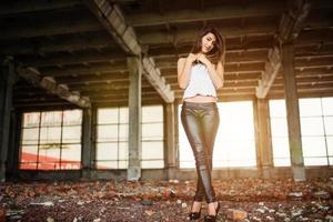 retrato de una joven morena linda con pantalones de cuero negro y blusa blanca posada en un lugar abandonado. foto