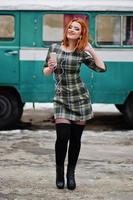 jovencita de pelo rojo con teléfono móvil y auriculares, vestida con un fondo de vestido a cuadros antiguo autobús minivan turquesa vintage. foto