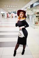 Fashion red haired girl wear on black dress and red hat with ladies backpack posed at trade shopping center. Photo toned style Instagram filters.