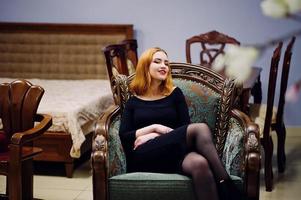 Fashion red haired girl wear on black dress with bright make up sitting on royal armchair. photo