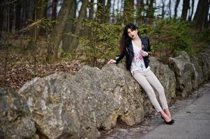 Brunette gypsy girl posed against stones on park. Model wear on leather jacket and t-shirt with ornament, pants and shoes with high heels. photo