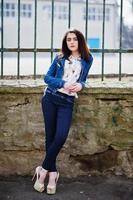 Young stylish brunette girl on shirt, pants, jeans jacket and high heels shoes, posed background iron fence. Street fashion model concept. photo