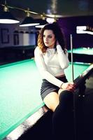 Young curly girl posed near billiard table. Sexy model at black mini mini skirt play russian snooker. Play game and fun concept. photo