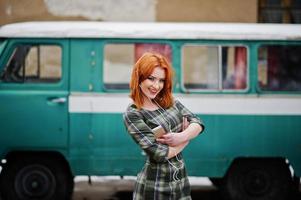 Young red haired girl lady with mobile cell phone and headphones, wearing on checked dress background old vintage turqoise minivan bus. photo