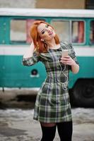 Young red haired girl lady with mobile cell phone and headphones, wearing on checked dress background old vintage turqoise minivan bus. photo