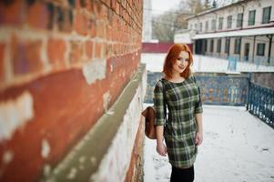 An outdoor portrait of a young pretty girl with red hair wearing checkered dress with girly backpacks standing on the brick wall background in winter day.