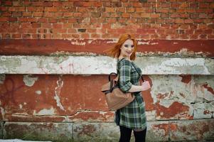 An outdoor portrait of a young pretty girl with red hair wearing checkered dress with girly backpacks standing on the brick wall background.