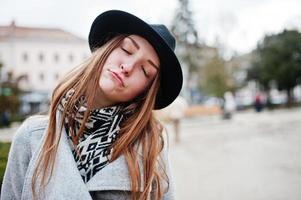 Cierra el retrato de una joven modelo divertida con un abrigo gris y un sombrero negro con los ojos cerrados y soñando en la calle de la ciudad. foto