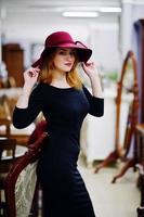 Portrait of fashion red haired girl on red hat and black dress with bright make up posed near table and chair salon. photo